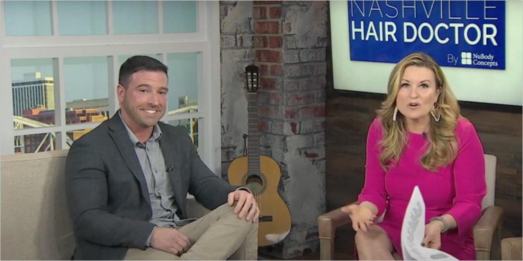 A man and a woman sitting down in front of a Nashville Hair Doctor sign.